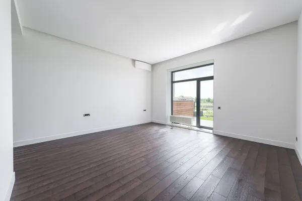 דירה ריקה עם רצפת פרקט כהה וחלון גדול שדרכו נכנס אור יום